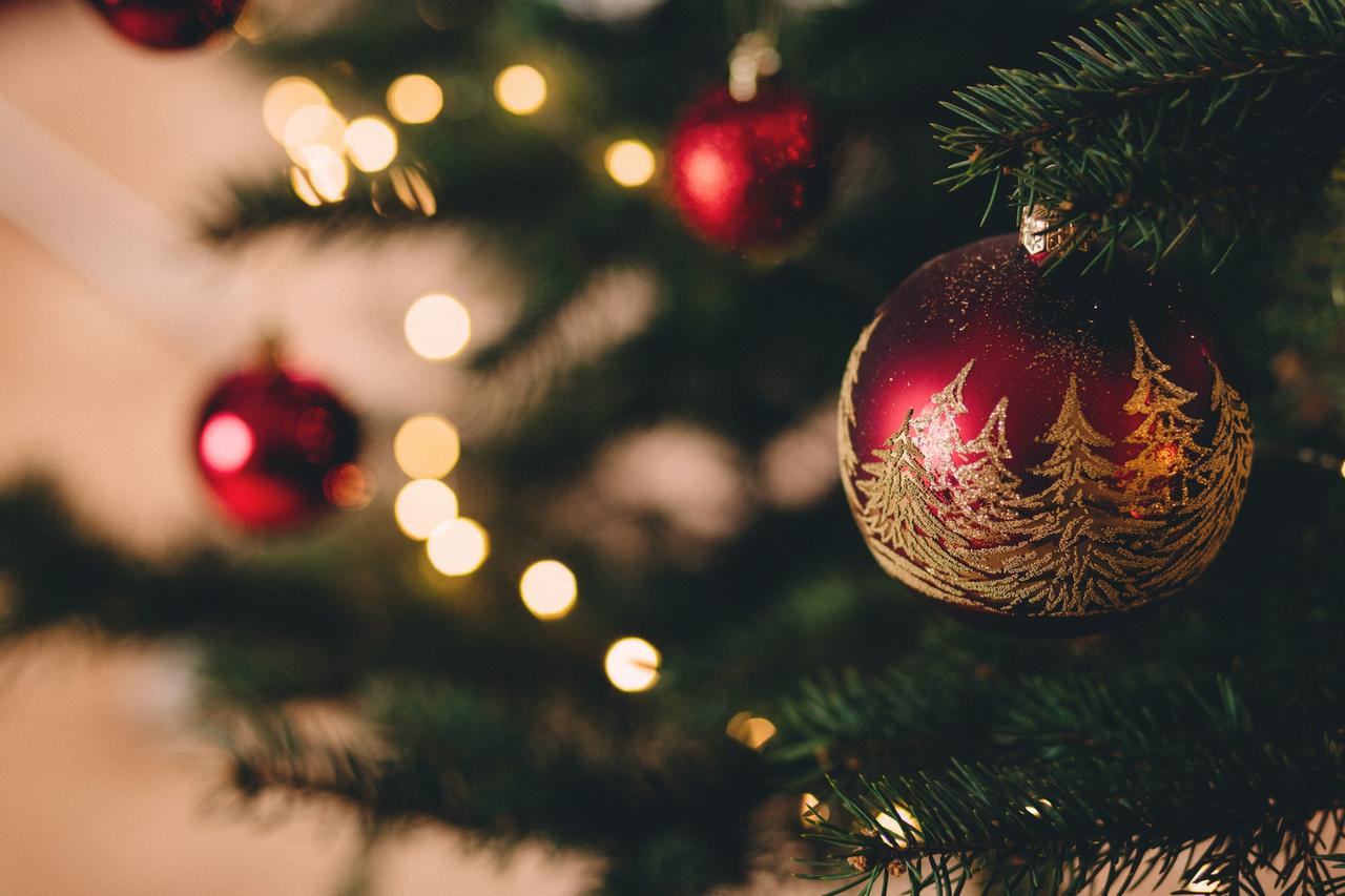 Im Vordergrund eine rote Christbaumkugel, die mit goldenen Tannen bemalt ist. Sie hängt an einem grünen Tannenbaum, der mit einer Lichterkette geschmückt wurde. Im Vordergrund eine rote Christbaumkugel, die mit goldenen Tannen bemalt ist. Sie hängt an einem grünen Tannenbaum, der mit einer Lichterkette geschmückt wurde.