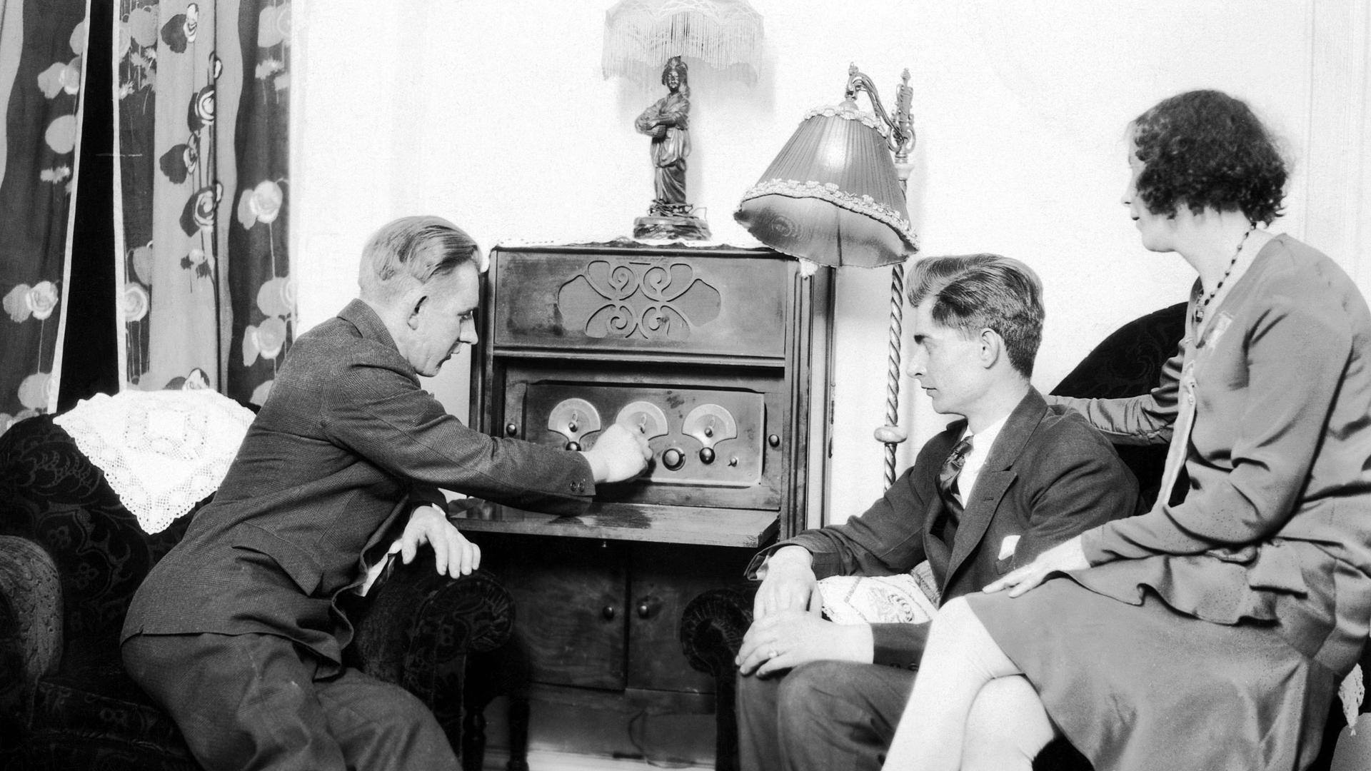 Zwei Männer und eine Frau sitzen in den 1920er Jahren in einem Wohnzimmer vor einem Radiogerät, dass einer der Männer gerade bedient. Zwei Männer und eine Frau sitzen in den 1920er Jahren in einem Wohnzimmer vor einem Radiogerät, dass einer der Männer gerade bedient.