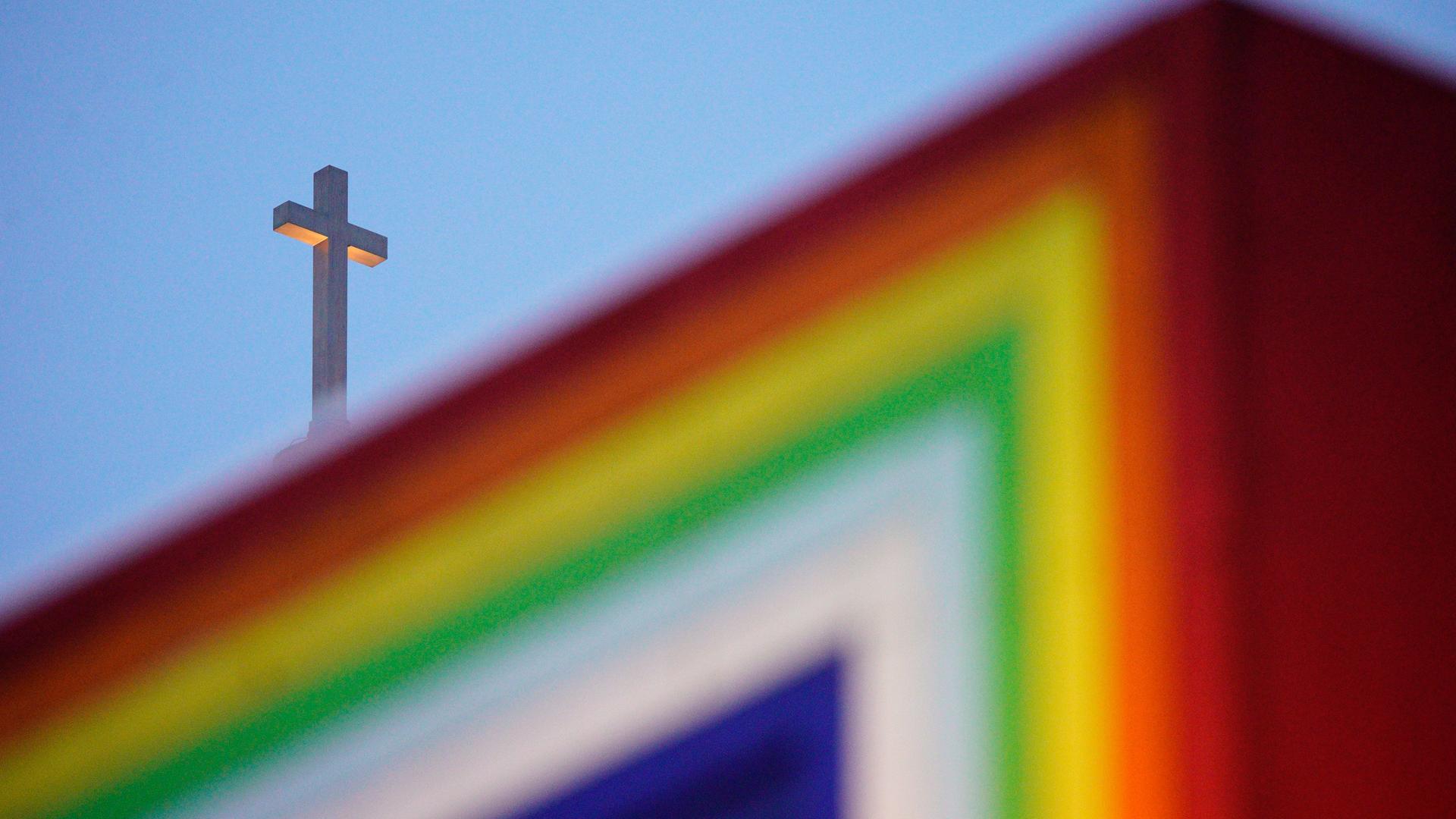 Hinter einer regenbogenfarbenen Skulptur ist ein Kreuz zu sehen. Hinter einer regenbogenfarbenen Skulptur ist ein Kreuz zu sehen.