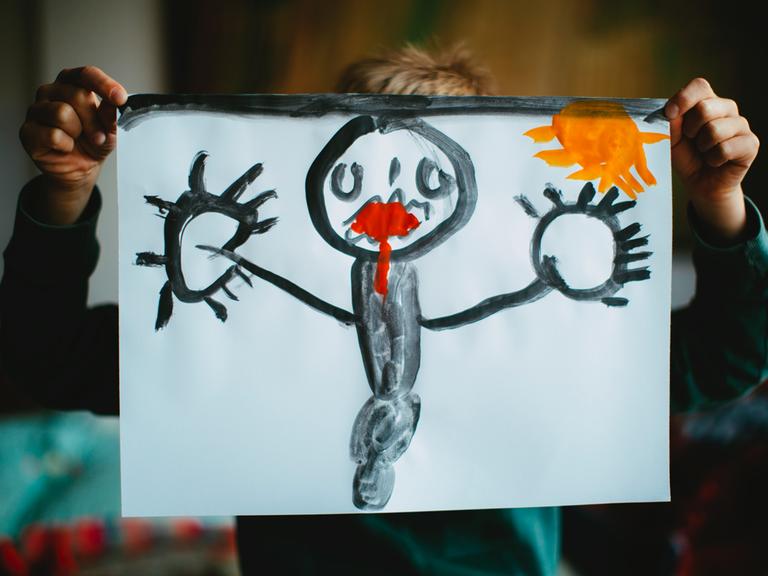 Ein Kind hält ein selbstgemaltes Bild vor seinen Kopf. Dem Mensch auf dem Bild läuft rote Farbe aus dem Mund.