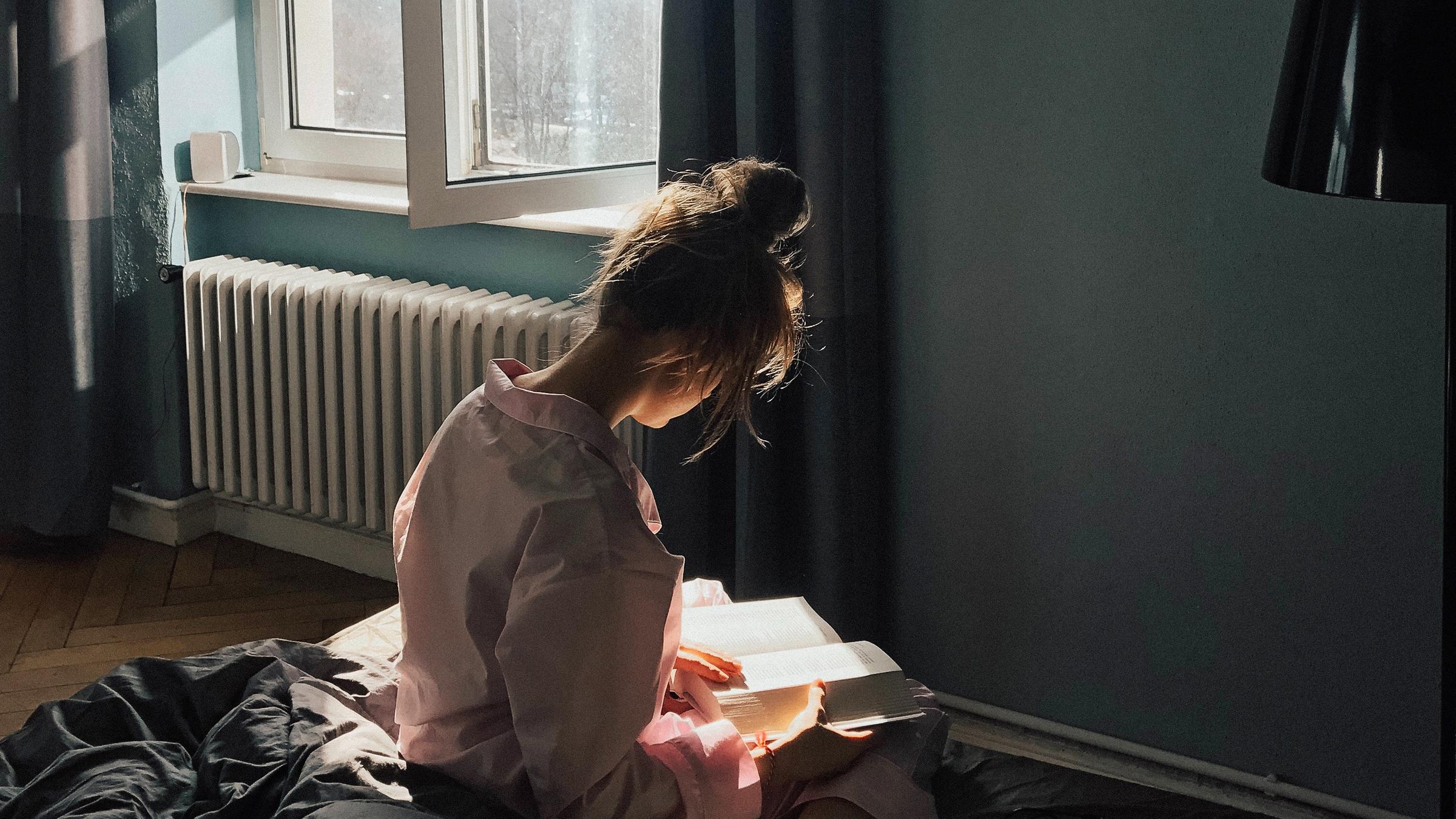 Eine junge Frau sitzt mit dem Rücken zum Betrachter vor dem Fenster auf dem Bett und hält ein geöffnetes Buch in den Händen.