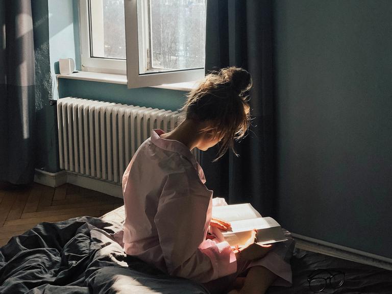 Eine junge Frau sitzt mit dem Rücken zum Betrachter vor dem Fenster auf dem Bett und hält ein geöffnetes Buch in den Händen.