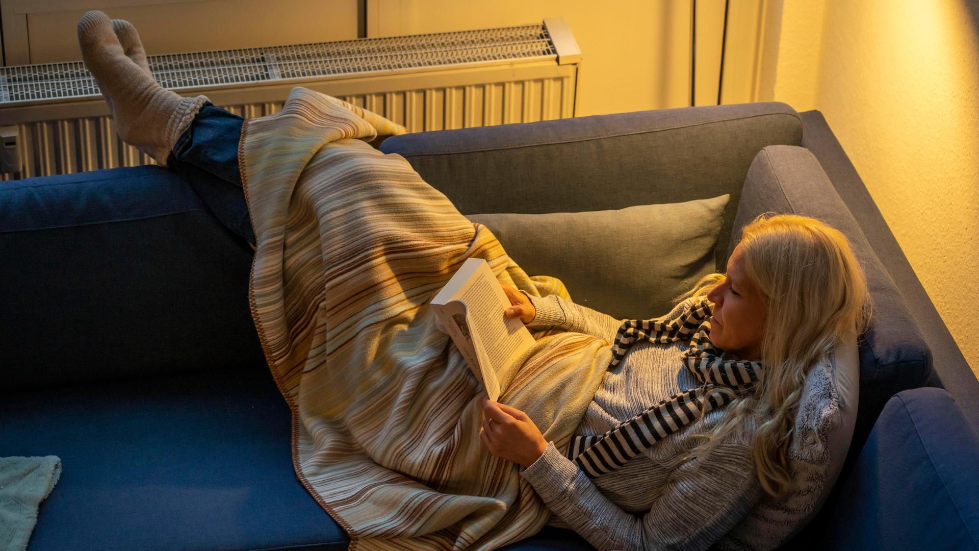 Eine Frau liegt warm angezogen mit Schal, dicken Socken und Decke bei Kerzenlicht auf einer Couch, dahinter ist ein Heizkörper zu sehen Eine Frau liegt warm angezogen mit Schal, dicken Socken und Decke bei Kerzenlicht auf einer Couch, dahinter ist ein Heizkörper zu sehen