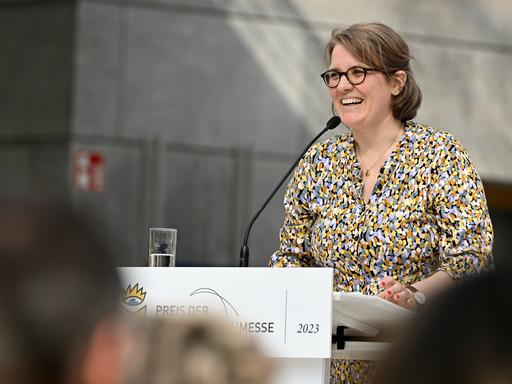 Johanna Schwering steht am Rednerpult auf der Leipziger Buchmesse. Sie trägt Brille und eine bunte Bluse. Im Vordergrund sind unscharf weitere Besucher der Messe zu sehen.