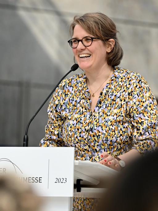 Johanna Schwering steht am Rednerpult auf der Leipziger Buchmesse. Sie trägt Brille und eine bunte Bluse. Im Vordergrund sind unscharf weitere Besucher der Messe zu sehen.