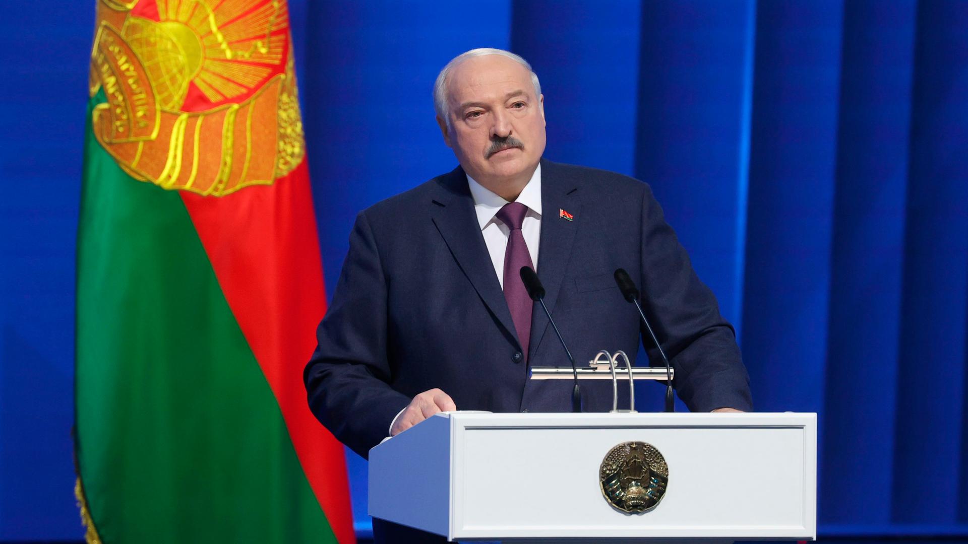 Der belarussische Präsident Lukaschenko steht hinter einem Rednerpult. Hinter ihm ein blauer Vorhang und die belarussische Fahne.