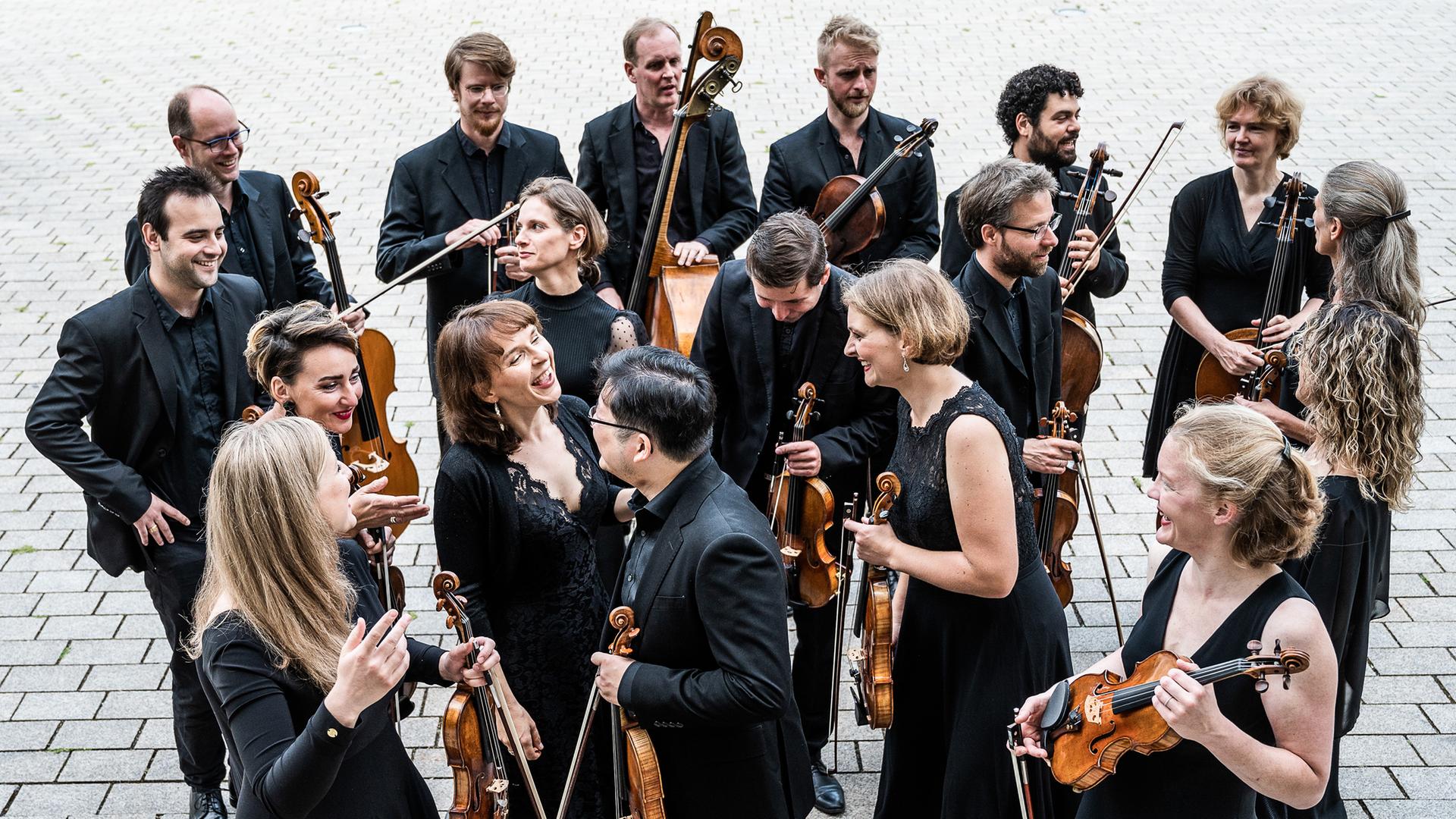 Musikerinnen und Musiker in schwarzer Konzertkleidung stehen in einer lockeren Gruppe und lachen einander an, während sie ihre Streichinstrumente in den Händen halten. Musikerinnen und Musiker in schwarzer Konzertkleidung stehen in einer lockeren Gruppe und lachen einander an, während sie ihre Streichinstrumente in den Händen halten.