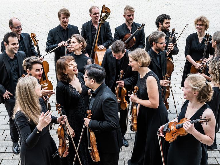Musikerinnen und Musiker in schwarzer Konzertkleidung stehen in einer lockeren Gruppe und lachen einander an, während sie ihre Streichinstrumente in den Händen halten.