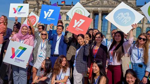 Teilnehmer des Jugendgipfels Y7 stehen vor dem Reichstagsgebäude in Berlin. Sie halten zum Teil Schilder in die Luft mit dem Logo des Gipfels und anderen Symbolen.