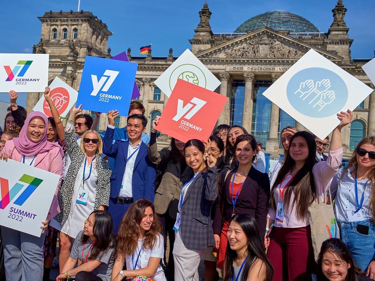 Teilnehmer des Jugendgipfels Y7 stehen vor dem Reichstagsgebäude in Berlin. Sie halten zum Teil Schilder in die Luft mit dem Logo des Gipfels und anderen Symbolen.