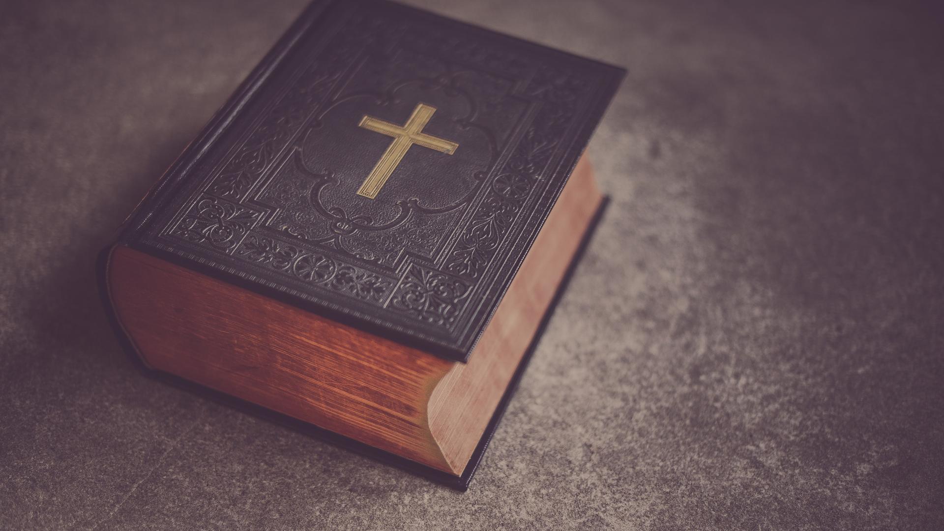 Eine schwere, dunkle Bibel mit einem goldenen Kreuz auf dem Deckel liegt auf einem Tisch. Eine schwere, dunkle Bibel mit einem goldenen Kreuz auf dem Deckel liegt auf einem Tisch.