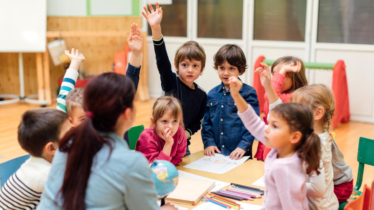Eine Kindergartenerzieherin unterrichtet Vorschulkinder in Geografie. Ein Globus steht auf dem Tischmitte und die Kinder strecken ihre Arme weil sie sich zu Wort melden wollen.