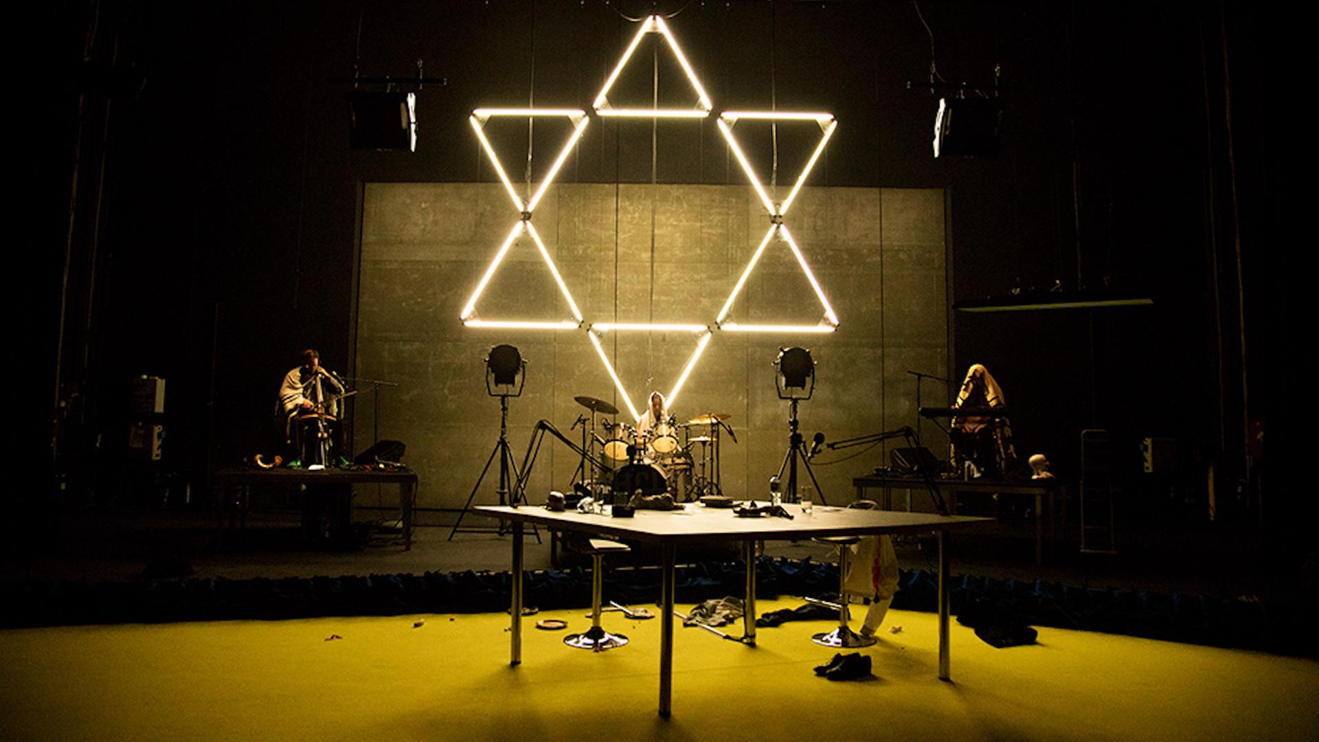 Hinter einer dreiköpfigen Band, die auf einer Bühne performt, hängt ein großer, aus Neonröhren zusammengesetzter Davidstern. Hinter einer dreiköpfigen Band, die auf einer Bühne performt, hängt ein großer, aus Neonröhren zusammengesetzter Davidstern.