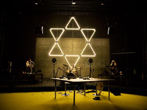 Hinter einer dreiköpfigen Band, die auf einer Bühne performt, hängt ein großer, aus Neonröhren zusammengesetzter Davidstern.