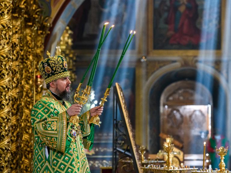 Das Oberhaupt der orthodoxen Kirche der Ukraine hält zwei Kerzenständer mit grünen Kerzen in seinen Händen. Im Bildhintergrund sind einzelne Sonnenstrahlen zu sehen, die von oben in die Kathedrale einfallen.