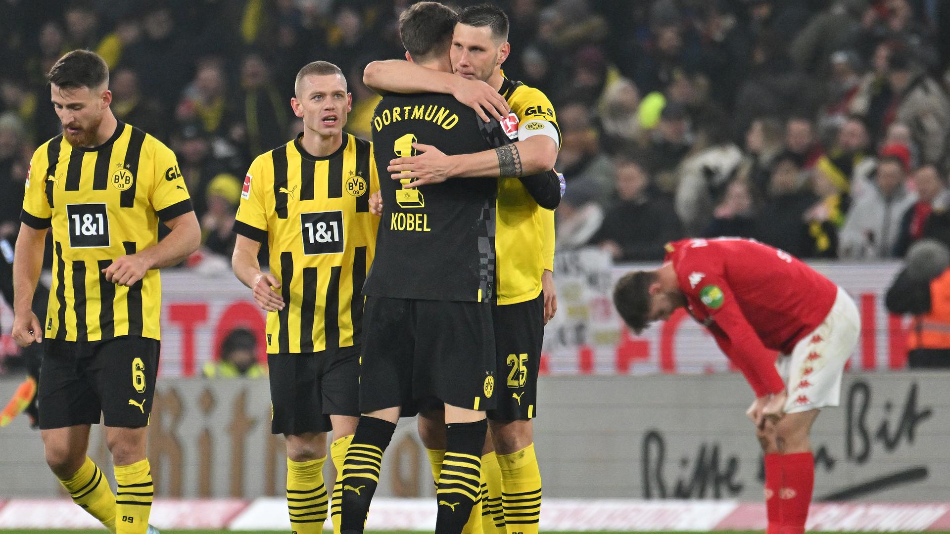 Spieler von Borussia Dortmund feiern nach dem Abpfiff ihren Sieg gegen Mainz und umarmen sich. Im Hintergrund ist ein Mainzer Spieler zu sehen, der sich mit frustriert herunterhängendem Kopf mit seinen Händen auf seine Knie aufstützt.