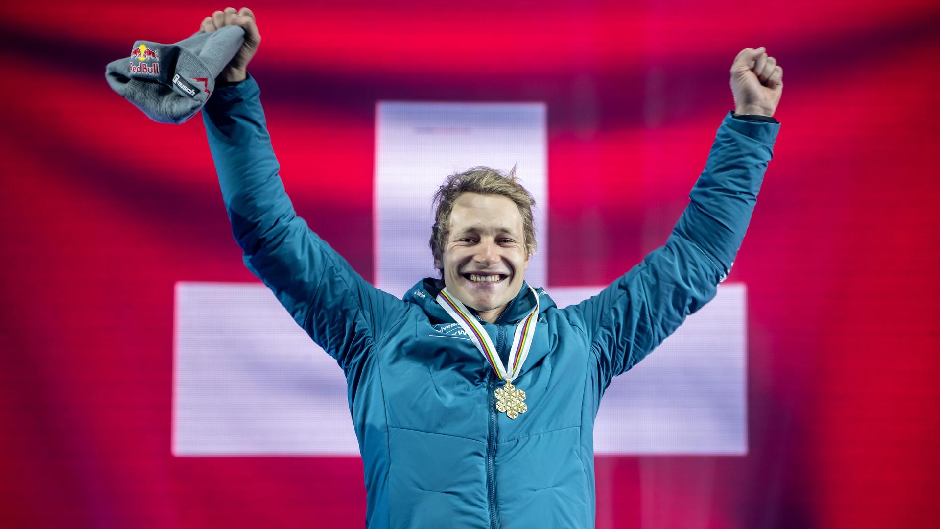 Ein junger, blonder Mann in blauer Winterjacke reißt jubelnd seine Arme hoch und strahlt, um seinen Hals hängt eine Medaille in Form eines Eiskristalls. Im Hintergrund ist unscharf die Fahne der Schweiz zu sehen.
