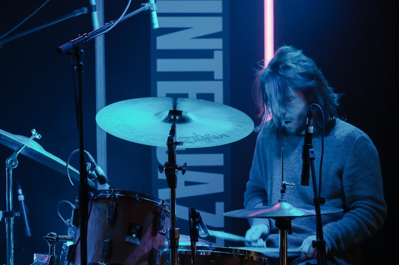 Ein Mann mit offenem, längerem Haar spielt, in blaues Schweinwerferlicht gehüllt, mit geschlossenen Augen Schlagzeug.