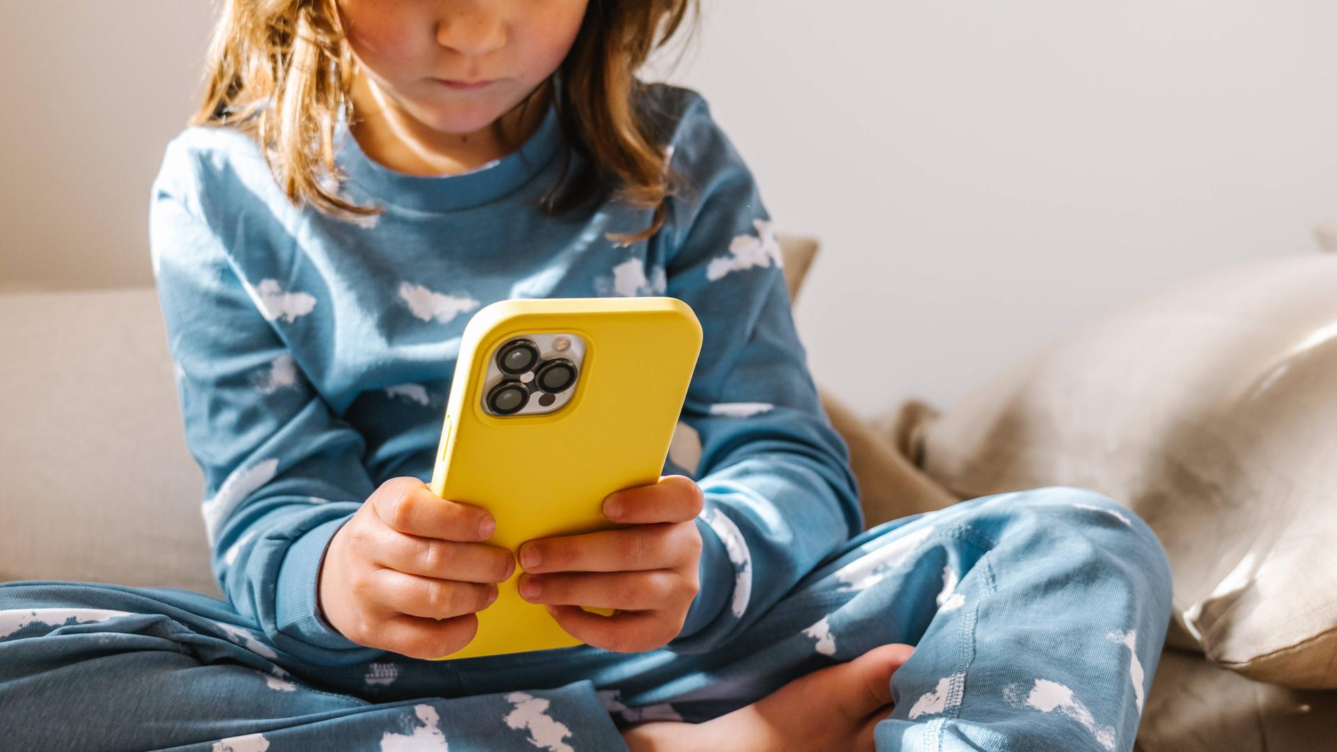 Ein Kleinkind im Schlafanzug sitzt auf einem Bett und klickt auf einem Smartphone in gelber Hülle. Ein Kleinkind im Schlafanzug sitzt auf einem Bett und klickt auf einem Smartphone in gelber Hülle.