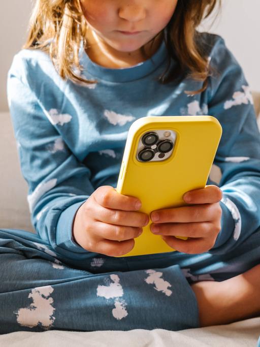 Ein Kleinkind im Schlafanzug sitzt auf einem Bett und klickt auf einem Smartphone in gelber Hülle.