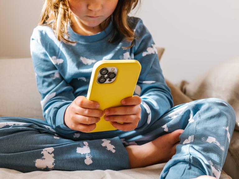 Ein Kleinkind im Schlafanzug sitzt auf einem Bett und klickt auf einem Smartphone in gelber Hülle.