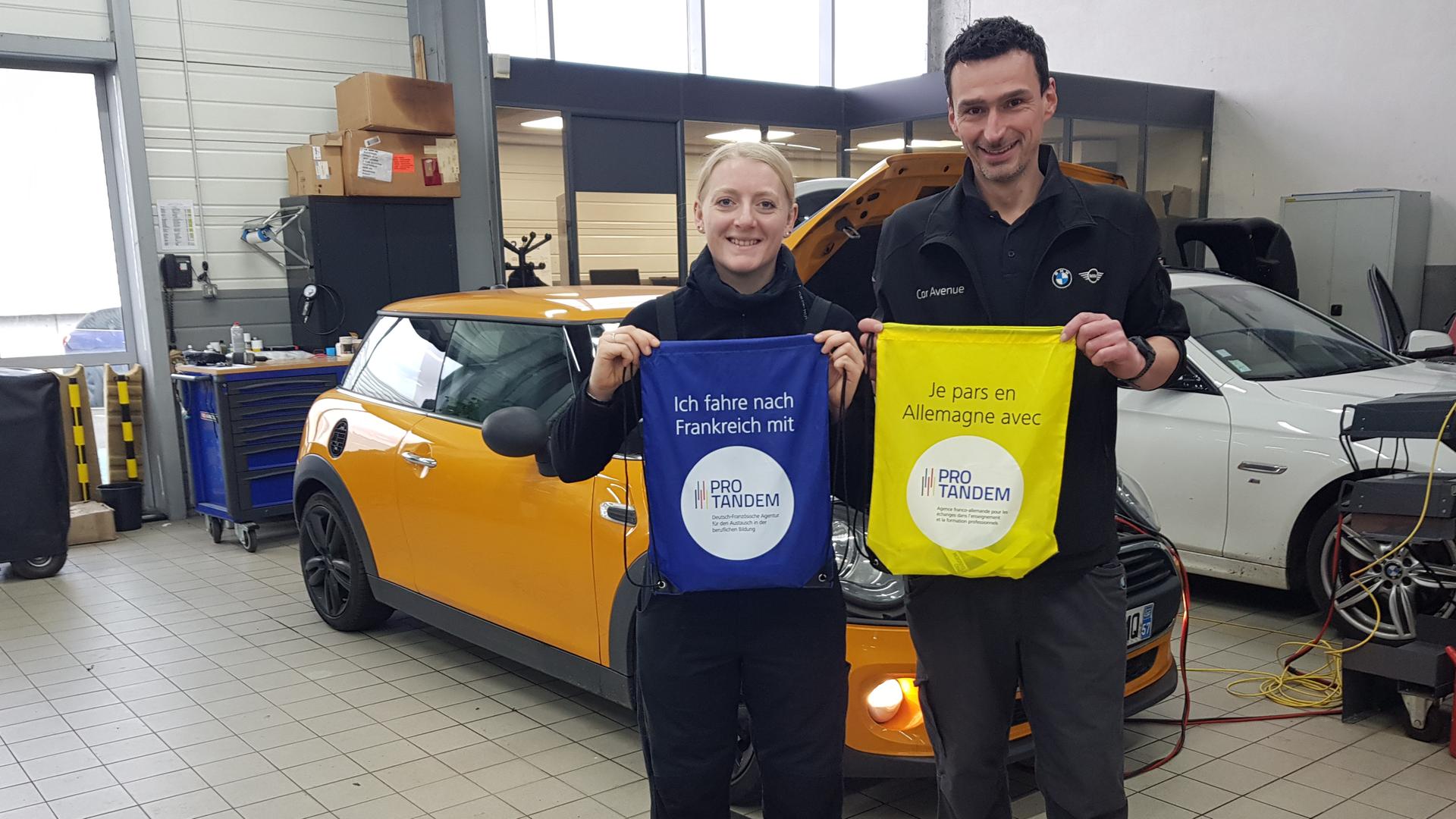 Eine junge, blonde Frau und ein etwas größerer Mann in dunklem Overall stehen in einer Kfz-Werkstatt. Die Frau hält einen blauen Gegenstand hoch mit dem Aufdruck "Ich fahre nach Frankreich mit Pro Tandem", der Mann einen gelben mit der Aufschrift "Je pars en Allemagne avec Pro Tandem". Eine junge, blonde Frau und ein etwas größerer Mann in dunklem Overall stehen in einer Kfz-Werkstatt. Die Frau hält einen blauen Gegenstand hoch mit dem Aufdruck "Ich fahre nach Frankreich mit Pro Tandem", der Mann einen gelben mit der Aufschrift "Je pars en Allemagne avec Pro Tandem".