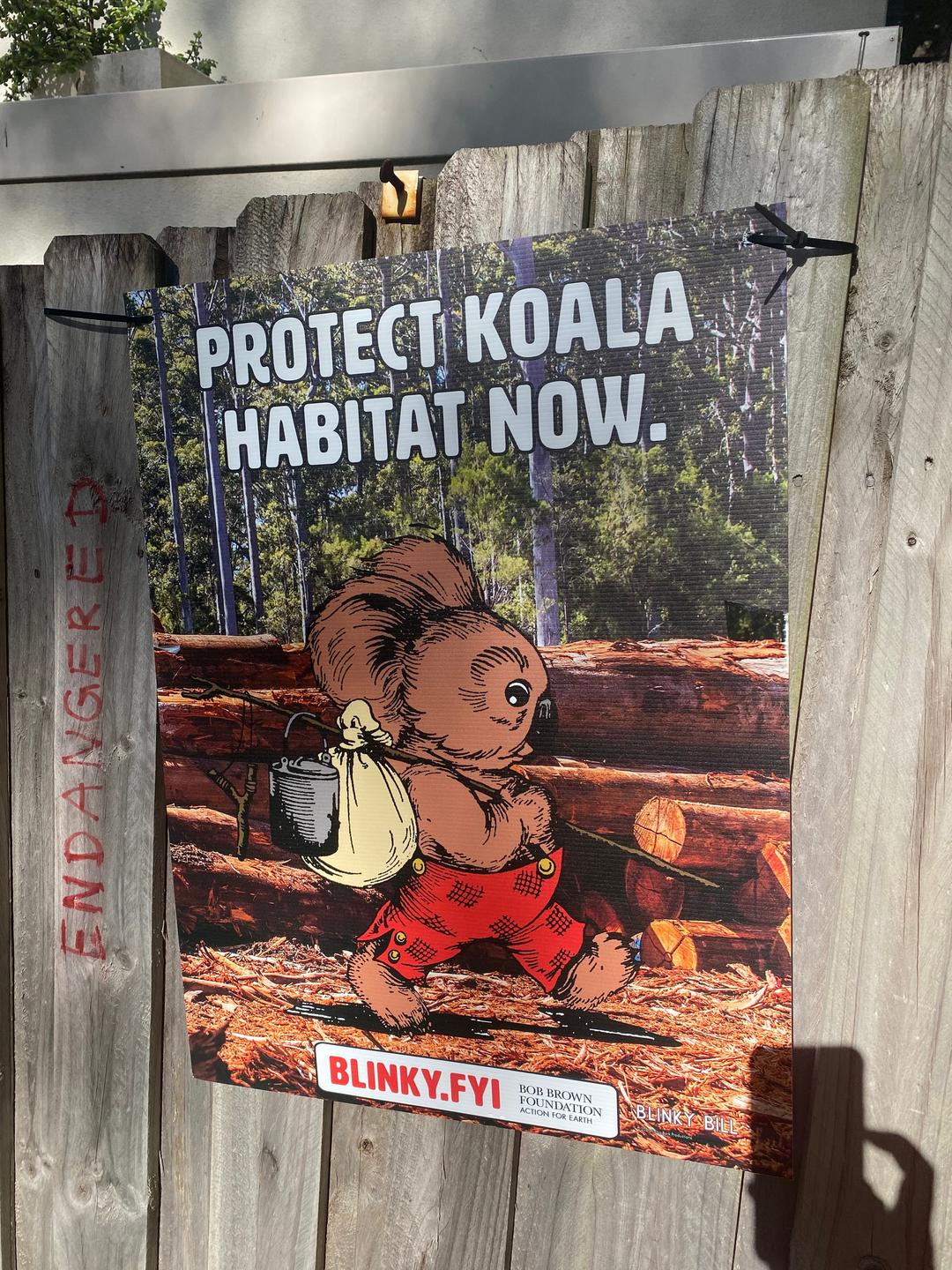 Auf einem Plakat an einem Holzzaun ist abgebildet: ein kleiner dunkelbrauner Koala, der mit Stock und Proviant durch den Wald marschiert.