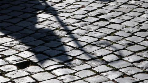 Der Schatten eines Bischofs mit Mitra und Bischofsstab ist auf einer gepflasterten Straße zu sehen.
