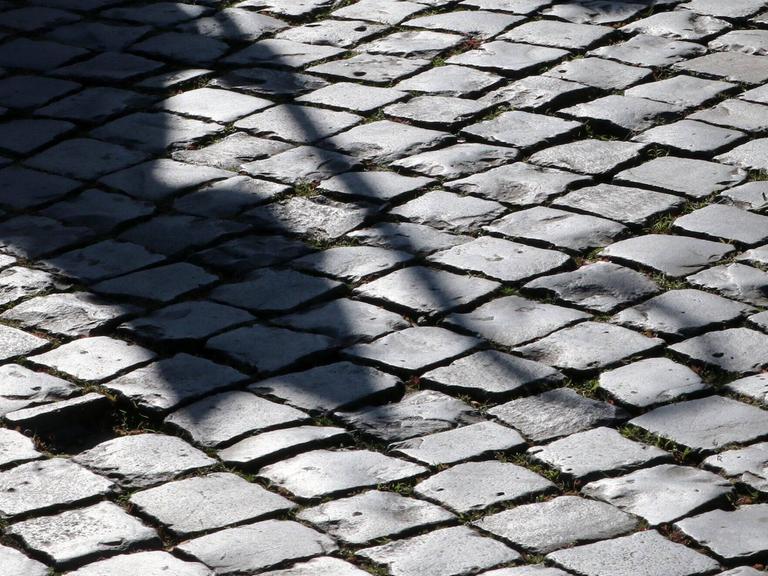 Der Schatten eines Bischofs mit Mitra und Bischofsstab ist auf einer gepflasterten Straße zu sehen.