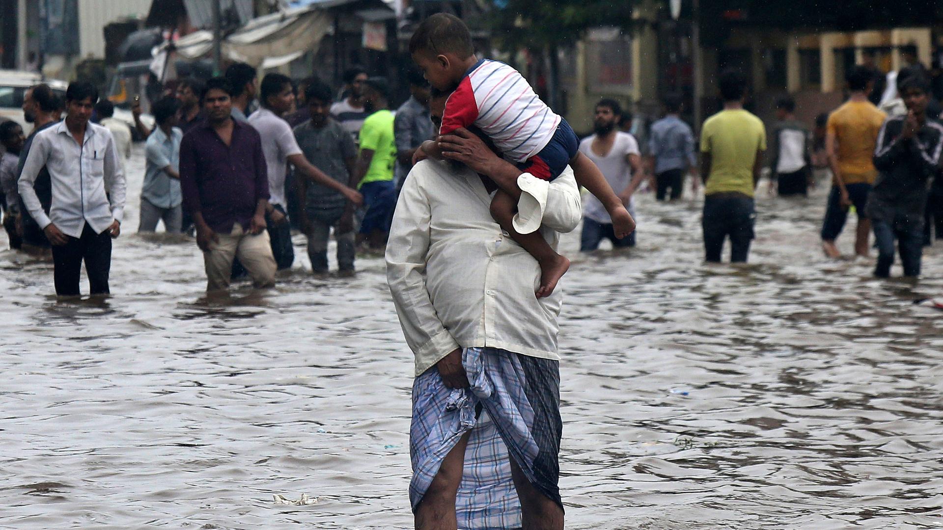 Menschen waten auf einer überfluteten Straße in Mumbai in Indien. Im Vordergrund trägt ein Mann ein Kleinkind.