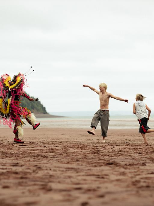 Eine Filmszene aus "Wildhood" zeigt drei Personen an einem sonst menschenleeren Meeresstrand. Zu sehen sind ein Mann in indigener Kostümierung und zwei Jungen beim Tanzen.