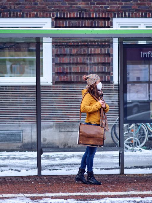 Eine Frau mit Mund-Nasen-Schutz steht an einer Bushaltestelle im Schnee.