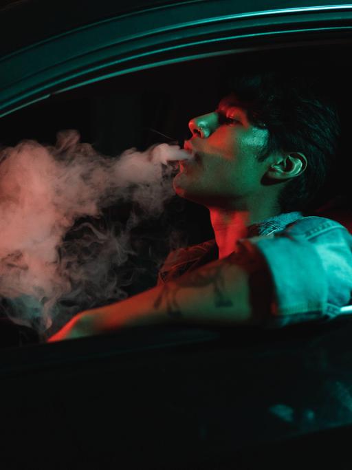 Ein junger Mann sitzt nachts in einem Auto und raucht.