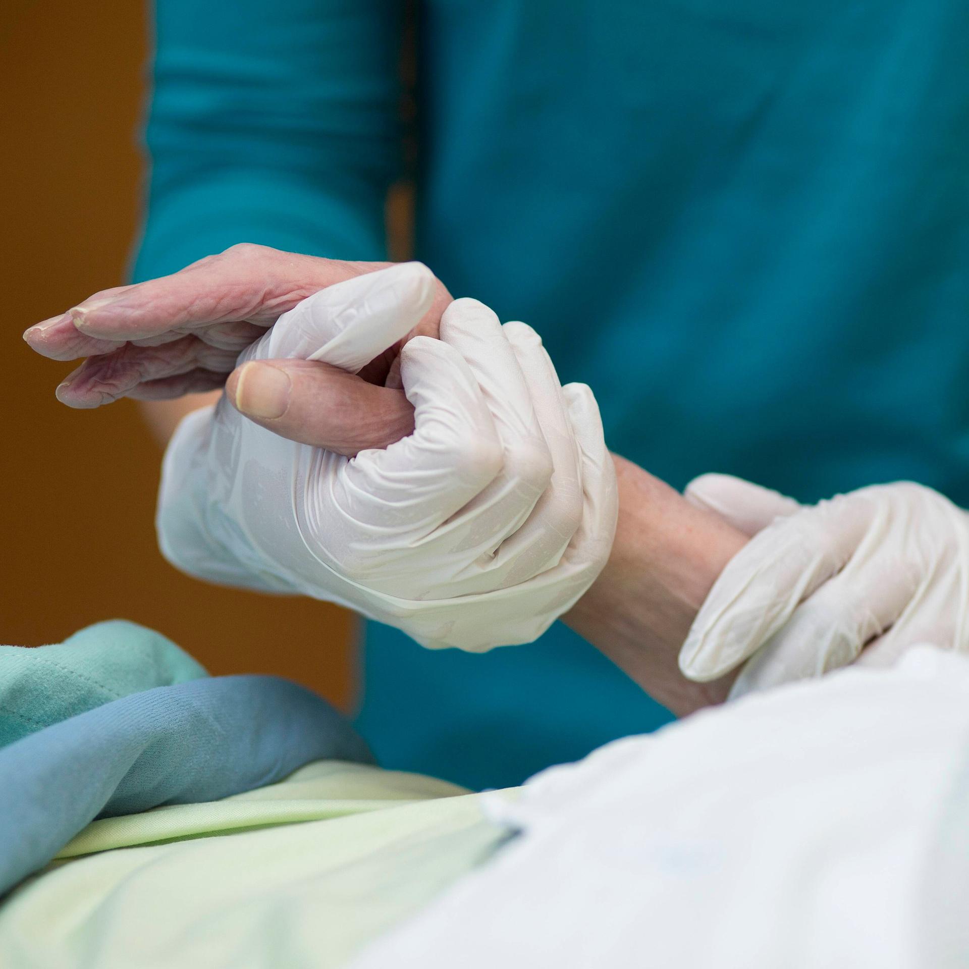 Eine junge Hand mit Hygienehandschuh hält eine alte Hand in einem Pflegebett.