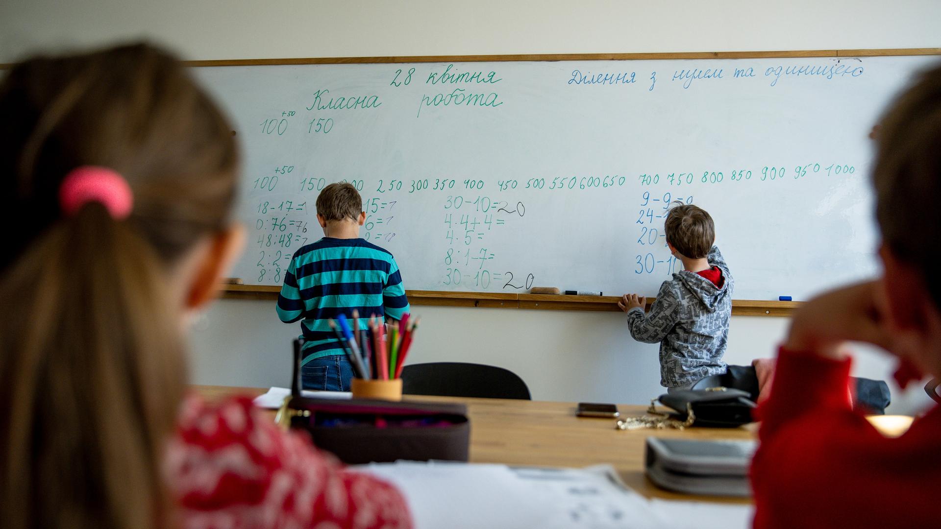 An einer Tafel die mit kyrillischen Buchstaben und mit Zahlen beschriftet sind, stehen zwei Kinder, die von hinten zu sehen sind. Im Vordergrund sind zwei weitere Kinder von hinten an einem Tisch zu sehen. An einer Tafel die mit kyrillischen Buchstaben und mit Zahlen beschriftet sind, stehen zwei Kinder, die von hinten zu sehen sind. Im Vordergrund sind zwei weitere Kinder von hinten an einem Tisch zu sehen.