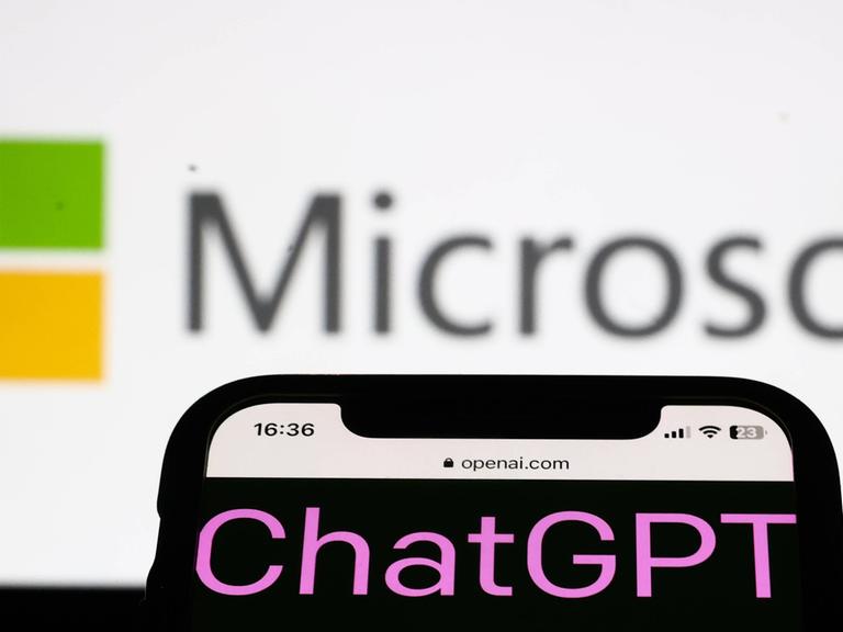 Ein Smartphone mit dem Wort ChatGPT auf dem Screen wird auf einen Bildschirm gerichtet, auf dem das Logo und der Schiftzug der Firma Microsoft zu sehen sind.