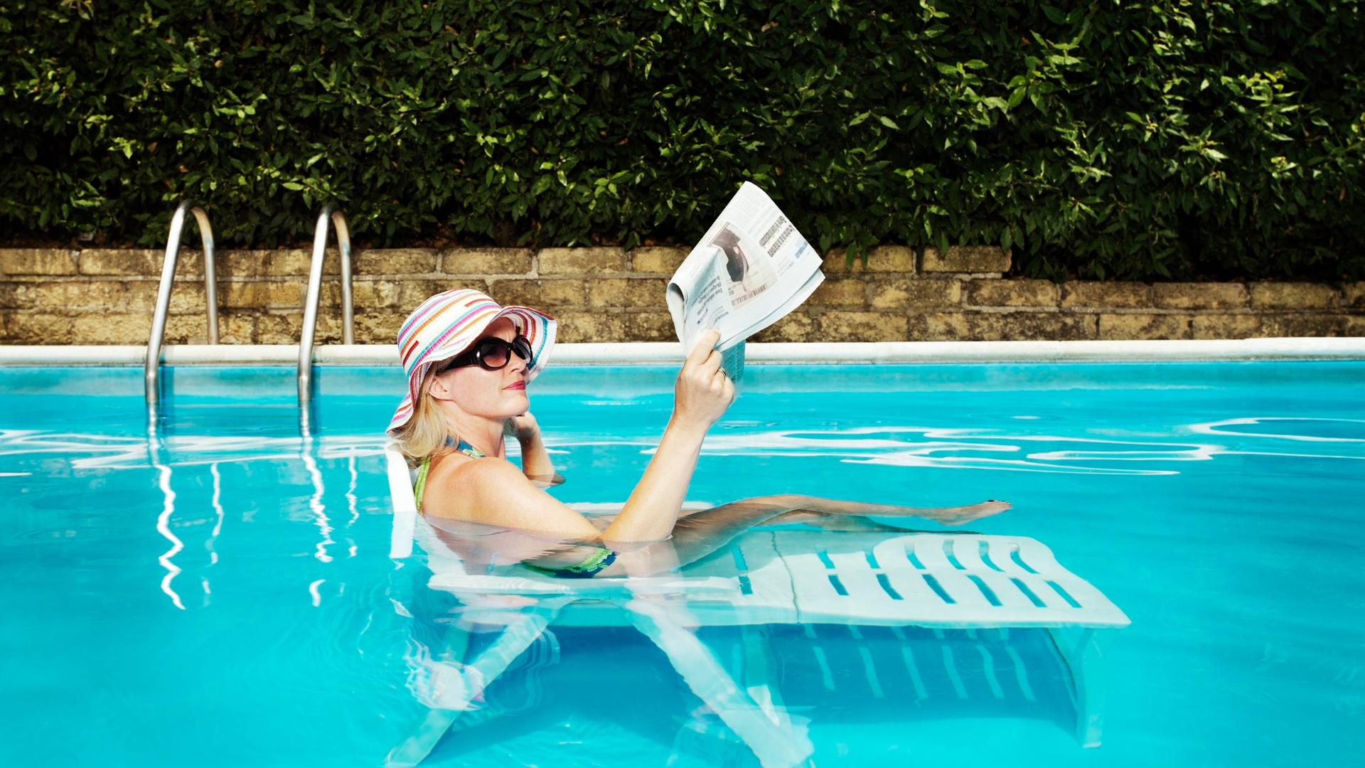 Eine Frau mit einem Sonnenhut liegt auf einer Sonneliege in einem gefüllten Pool und liest Zeitung. Eine Frau mit einem Sonnenhut liegt auf einer Sonneliege in einem gefüllten Pool und liest Zeitung.