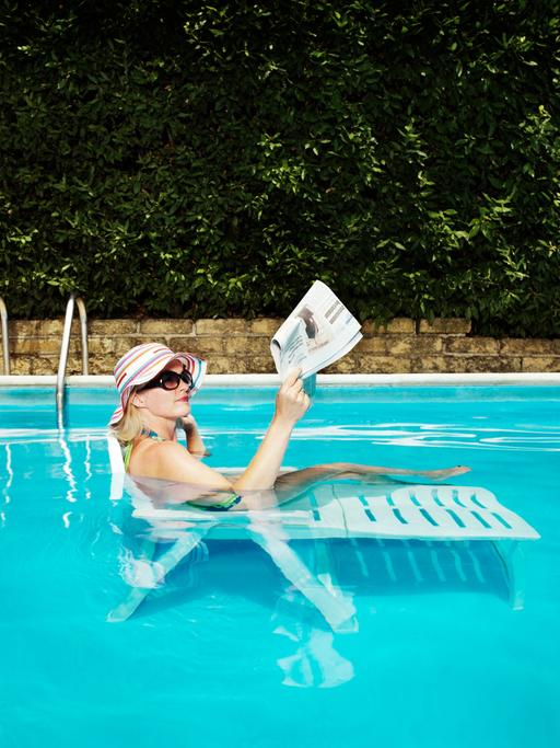 Eine Frau mit einem Sonnenhut liegt auf einer Sonneliege in einem gefüllten Pool und liest Zeitung.