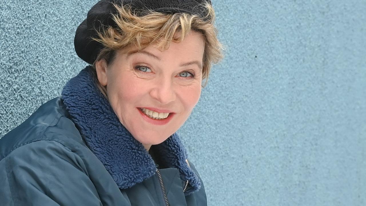 Eine Frau mit schwarzer Kappe, unter der blonde Locken hervorblitzen, sitzt vor einer blaugrauen Wand in der Hocke und lächelt in die Kamera. Eine Frau mit schwarzer Kappe, unter der blonde Locken hervorblitzen, sitzt vor einer blaugrauen Wand in der Hocke und lächelt in die Kamera.
