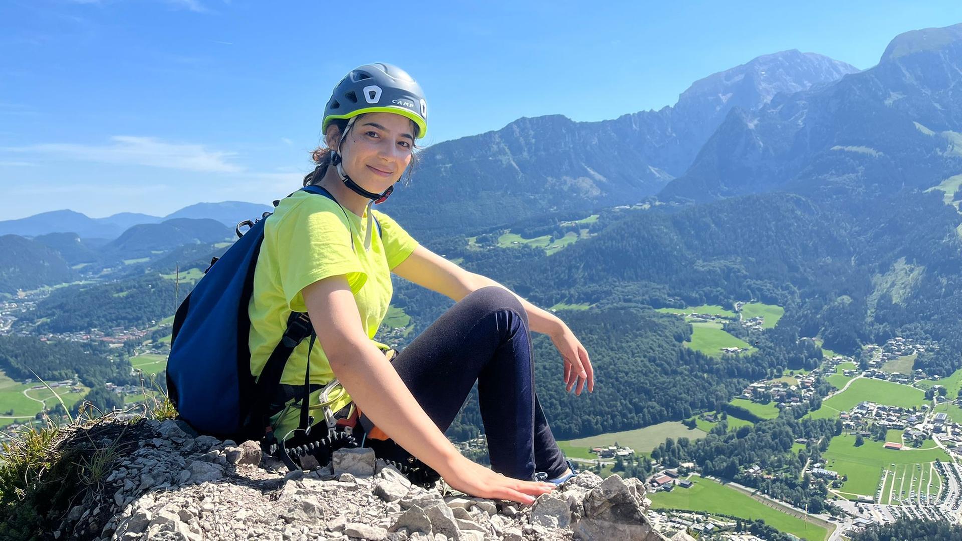 Eine Frau mit Helm und Rucksack sitzt auf einem Felsen, im Hintergrund ist ein Bergmassiv zu sehen.