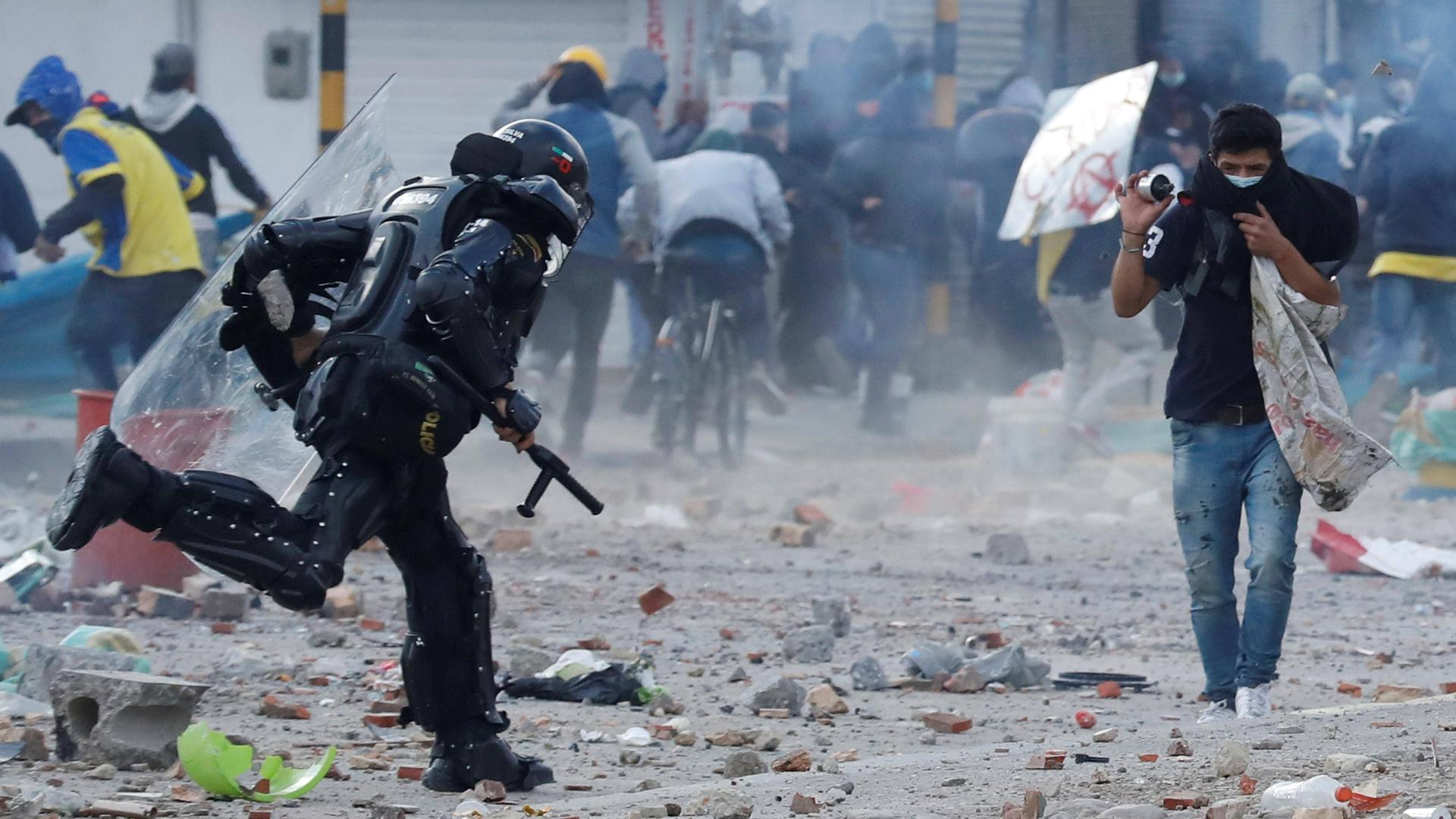 Ein Polizist in voller Schutzausrüstung und mit Schlagstock rennt auf einen Demonstranten zu. Im Hintergrund laufen viele Demonstranten weg. Ein Polizist in voller Schutzausrüstung und mit Schlagstock rennt auf einen Demonstranten zu. Im Hintergrund laufen viele Demonstranten weg.
