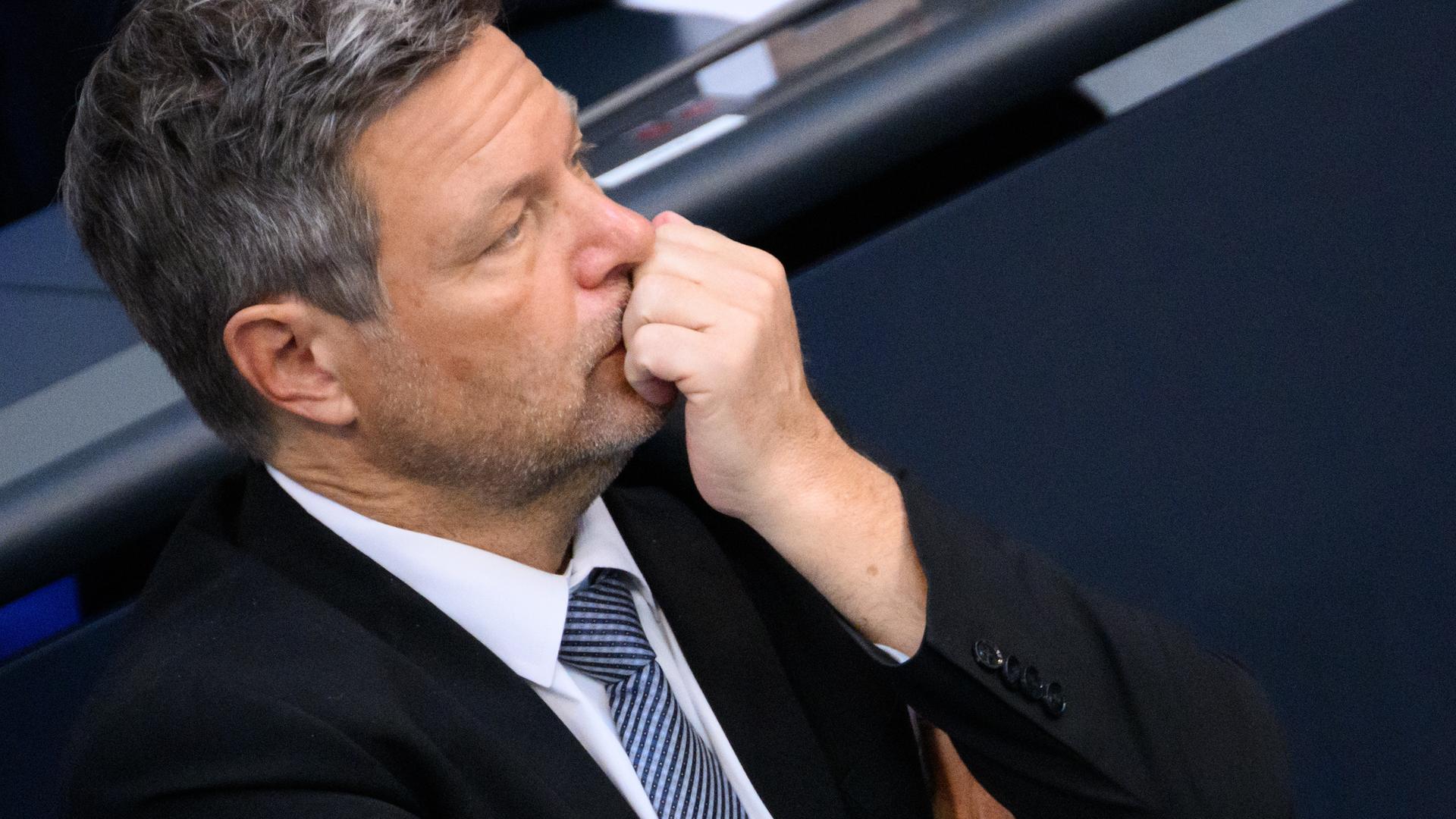 Bundeswirtschaftsminister Robert Habeck (re., Bündnis 90/Die Grünen) sitzt in schwarzem Anzung und weissem Hemd im Bundestag. Er hält sich die linke Faust vor das Gesicht und guckt nach links.