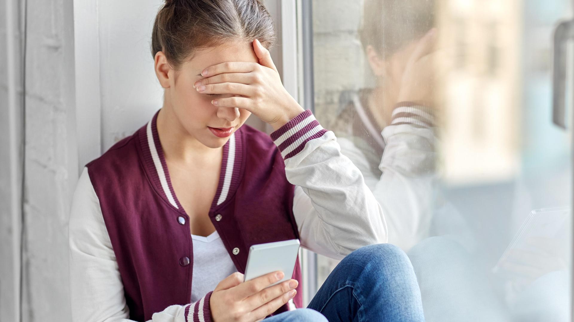 Eine junge Person sitzt alleine in einer Ecke und schaut auf ein Smartphone. Eine junge Person sitzt alleine in einer Ecke und schaut auf ein Smartphone.
