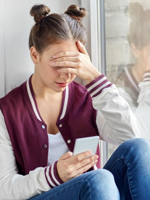 Eine junge Person sitzt alleine in einer Ecke und schaut auf ein Smartphone. 