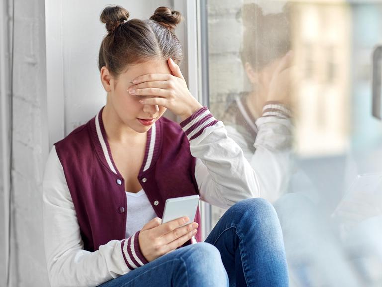 Eine junge Person sitzt alleine in einer Ecke und schaut auf ein Smartphone. 