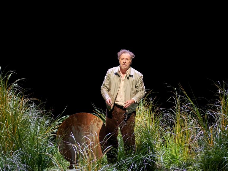 Ein Mann mit grauem, zerzaustem Haar steht auf einer Bühne. Er ist umgeben von hohem Gras und guck verwirrt. Er träge eine beige Jacke. 