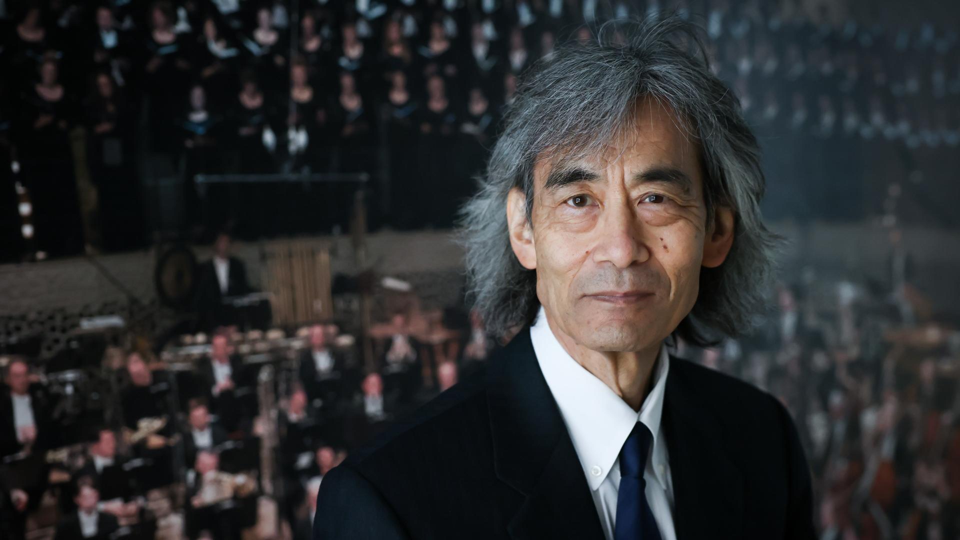 Ein Mann mit ergrauten, längeren Haaren steht vor enem Poster eines Konzerts aus der Elbphilharomie, auf dem viele Musiker zu sehen sind. Er trägt einen dunklen Anzug, eine dunkle Krawatte und ein helles Hemd.