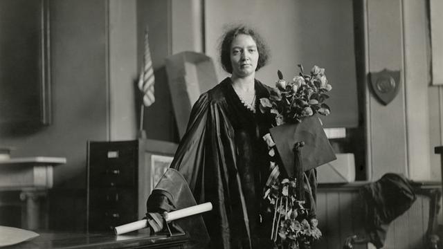 Die Physikerin Irene Joliot-Curie in akademischen Insignien: Sie trägt einen festlichen Talar, hält einen großen Blumenstrauß in der linken Hand und eine Schriftrolle in der rechten.