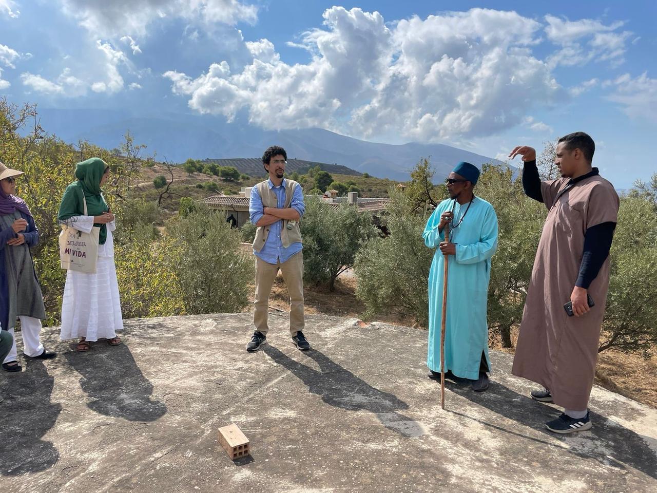Eine vierköpfige Gruppe steht auf einem von Bäumen umgebenen Platz und hört einem Vortragenden in traditionell muslimischer Kleidung zu. Eine vierköpfige Gruppe steht auf einem von Bäumen umgebenen Platz und hört einem Vortragenden in traditionell muslimischer Kleidung zu.
