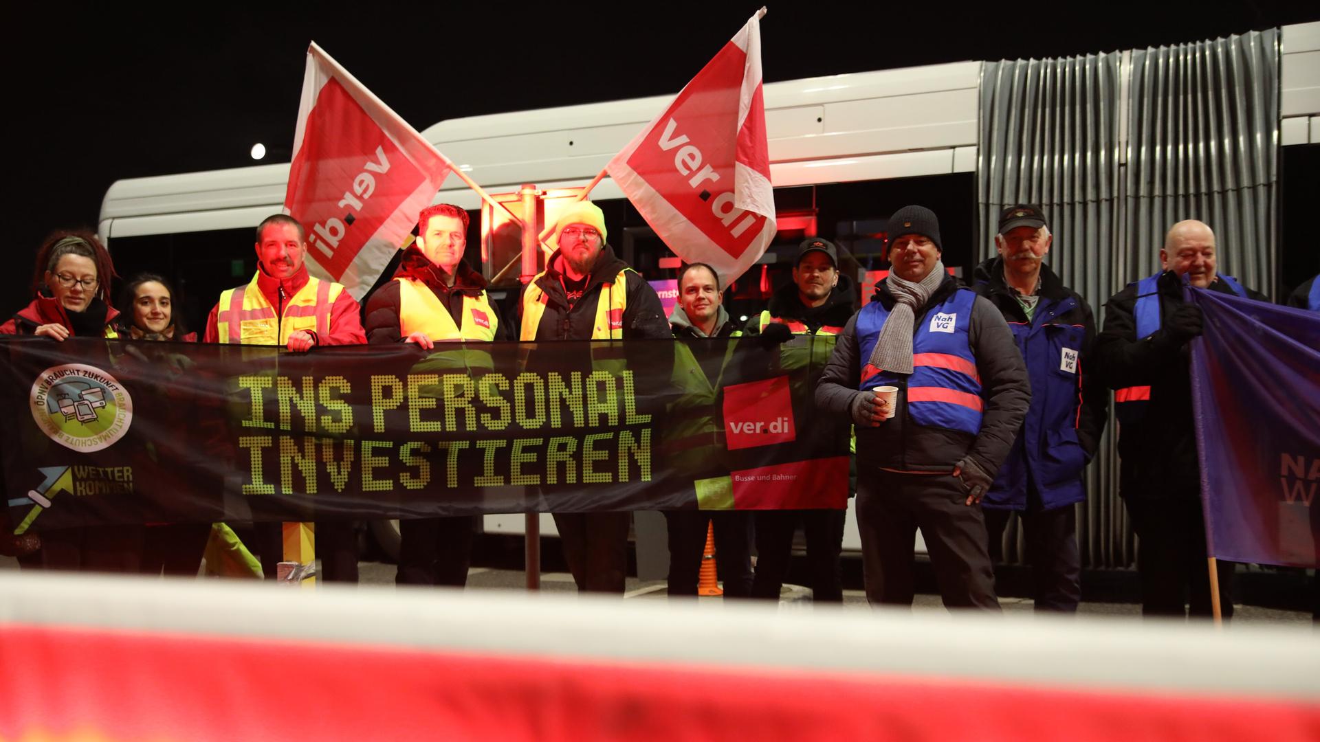 Eine Gruppe Streikender steht in Warnwesten vor einem Bus und hält ein Banner in der Hand, auf dem steht "Ins Personal investieren". Eine Gruppe Streikender steht in Warnwesten vor einem Bus und hält ein Banner in der Hand, auf dem steht "Ins Personal investieren".